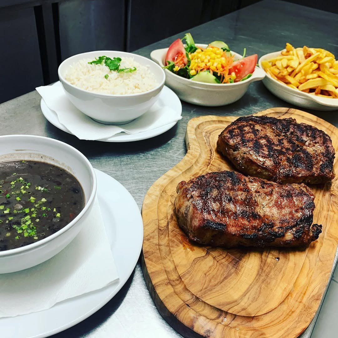 Dois bifes grelhados numa tábua de madeira, com arroz, feijão preto, salada e batatas fritas à volta
