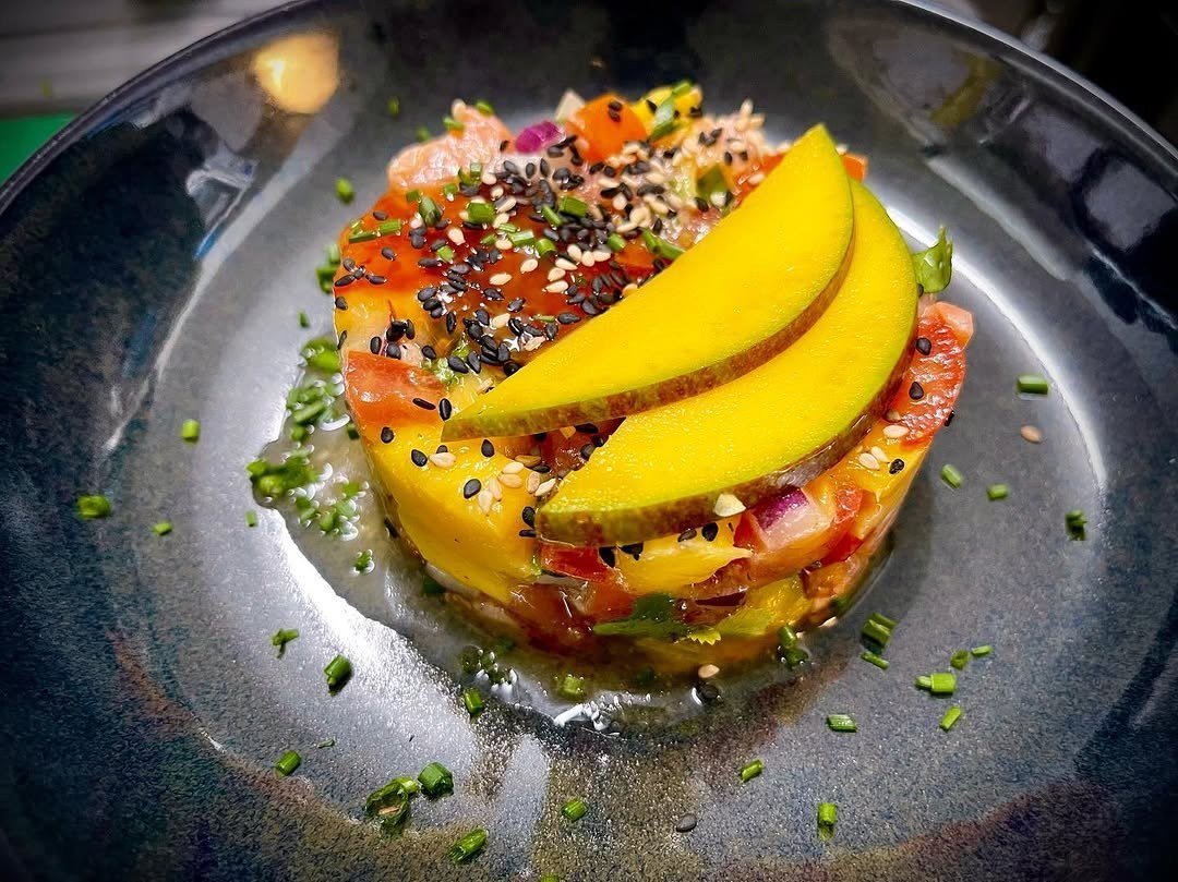 Tartar de salmão em torre com fatias de manga por cima e cebolinho à volta, num prato escuro