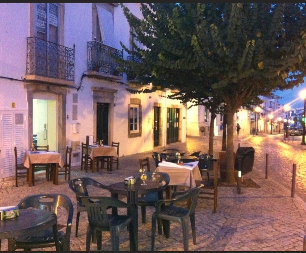 Esplanada do restaurante ao entardecer, com mesas e cadeiras numa praceta empedrada, junto a uma árvore e a casas brancas com varandas