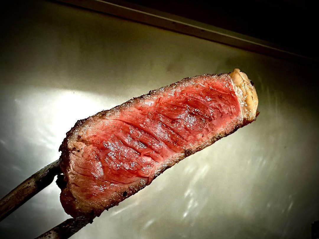 Bife grelhado espetado num garfo de churrasco, cortado ao meio e rosado por dentro, sobre fundo escuro